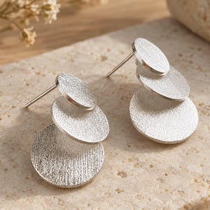 May include: A pair of silver-toned stud earrings, each featuring three graduated circular discs with a brushed, textured finish. The discs are arranged in a cascading vertical design, connected by a central post mechanism for secure attachment.