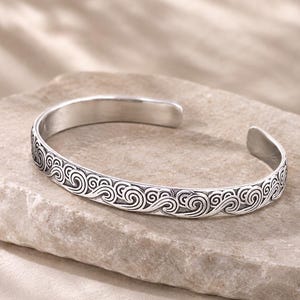 May include: A silver cuff bracelet featuring a continuous embossed pattern of stylized ocean waves. The metal has a polished finish on the interior and a darkened, oxidized recessed area within the wave design to highlight the details.