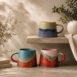 Puede incluir: Tres tazas de cer&aacute;mica con forma c&oacute;nica y un acabado de esmalte reactivo bicolor. Una taza presenta tonos azules y verdes, otra un dise&ntilde;o azul y rojo anaranjado, y la tercera muestra una combinaci&oacute;n de colores rosa y rojo con una base texturizada.