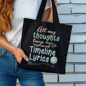 Op de afbeelding: Zwarte draagtas met de tekst "All my thoughts have been replaced with Timeline Lyrics" in een kleurrijk lettertype. De tas heeft een wereldbol-afbeelding en lange zwarte handvatten.