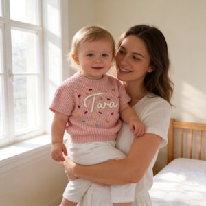 May include: A pink knit sweater with the name "Tara" embroidered in white script. The sweater has colorful embroidered accents and is worn by a child. The child is held by an adult in a white dress.