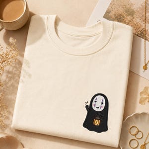 Op de afbeelding: Een crèmekleurig T-shirt met ronde hals en een kleine geborduurde afbeelding aan de rechteronderkant. Het borduursel toont een zwart, spookachtig figuur met een wit masker, paarse accenten en een klein geel lantaarntje in de hand.