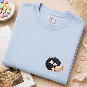 Op de afbeelding: Een lichtblauw sweatshirt met ronde hals en een geborduurd ontwerp van een klein, rond, zwart pluizig wezen met gesloten ogen dat een veelkleurig gestreept snoepje vasthoudt. Het label in de nek toont de tekst Comfort Colors en maat M-M.