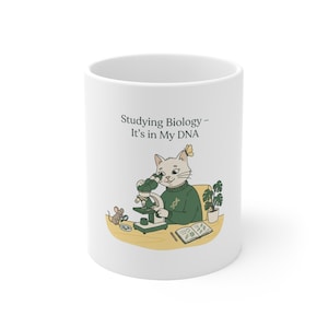 Peut inclure: Un mug en céramique blanc orné d'une illustration de chat portant un pull vert avec un brin d'ADN, observant à travers un microscope. Une petite souris avec une loupe se tient à côté. Le texte indique : Studying Biology - It's in My DNA.