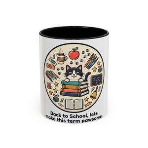 May include: A white ceramic mug with a black interior and rim, featuring a circular graphic of a black and white cat resting on a stack of books. Surrounding the cat are school supplies, an apple, and the text: Back to School, lets make this term pawsome.