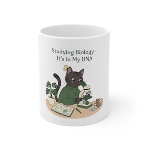 Peut inclure: Un mug en céramique blanche avec une illustration de chat tigré brun foncé portant un pull vert, observant à travers un microscope. Le texte indique "Studying Biology – It's in My DNA". Une petite souris avec une loupe est assise à côté.