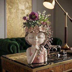 May include: A decorative planter shaped like a person with closed eyes and a finger held to their lips in a shushing gesture. The head is adorned with a crown of sculpted flowers and filled with a variety of purple, white, and green plants.