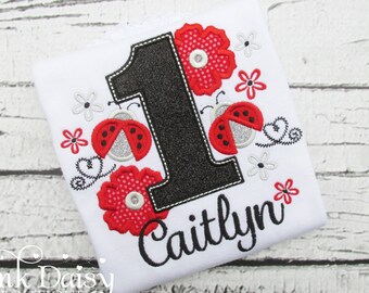 May include: White onesie with a black number one, red and black ladybugs, red flowers, and the name Caitlyn embroidered in black.