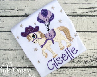 Puede incluir: Diseño bordado en morado y blanco de un caballo con un sombrero de vaquero y llevando globos morados. El caballo tiene una silla de montar con el número 6. El nombre Giselle está bordado debajo del caballo.