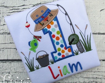 May include: A white onesie with a blue, green, orange, and red embroidered number one, a fishing pole, a bucket, a fish, and a fishing hat. The name Liam is embroidered in blue, green, red, and orange letters.