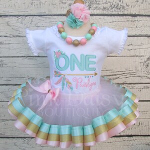 May include: A white onesie with the word "ONE" embroidered in teal with a gold arrow and feathers. The onesie is paired with a pink tulle tutu with gold, teal, and pink ribbon trim. The outfit is accessorized with a pink flower headband and a teal and gold beaded necklace.
