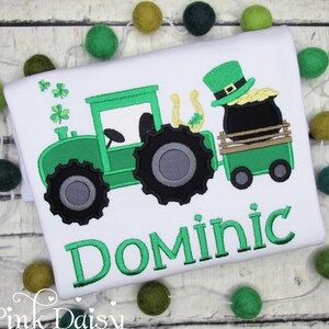 May include: Green tractor pulling a wagon with a pot of gold and a leprechaun. The wagon has a horseshoe on the side. The name Dominic is embroidered in green below the tractor.