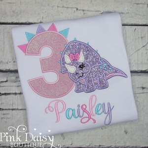 Puede incluir: Una camiseta blanca con un n&uacute;mero 3 brillante de color rosa y morado y un dinosaurio brillante de color morado con un lazo rosa. El nombre "Paisley" est&aacute; bordado en rosa, azul y morado.