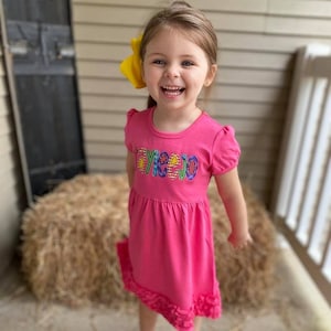 May include: A young girl wearing a pink dress with a colorful "Tayleejo" embroidery on the front. She is smiling and wearing a yellow bow in her hair.