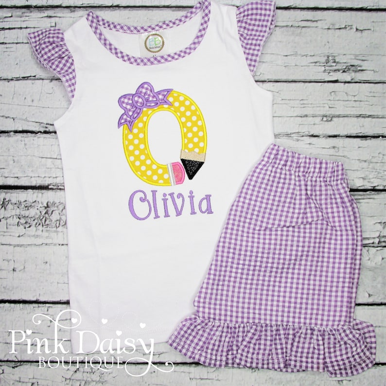 May include: A white sleeveless shirt with a purple and white gingham trim. The shirt has a yellow letter O with a pink bow and a pencil design. The name Olivia is embroidered in purple below the letter. The shirt is paired with a purple and white gingham skirt with a ruffle.