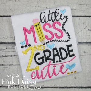May include: White t-shirt with embroidered text that reads "little miss 2nd grade cutie" with a pencil and hearts.