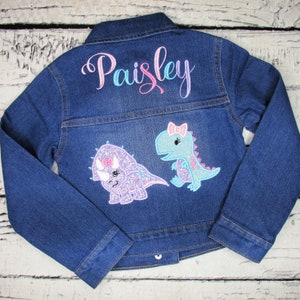 Puede incluir: Una chaqueta vaquera azul con el nombre "Paisley" bordado en la espalda. La chaqueta presenta dos dinosaurios bordados, uno morado y otro azul, con lazos rosas.