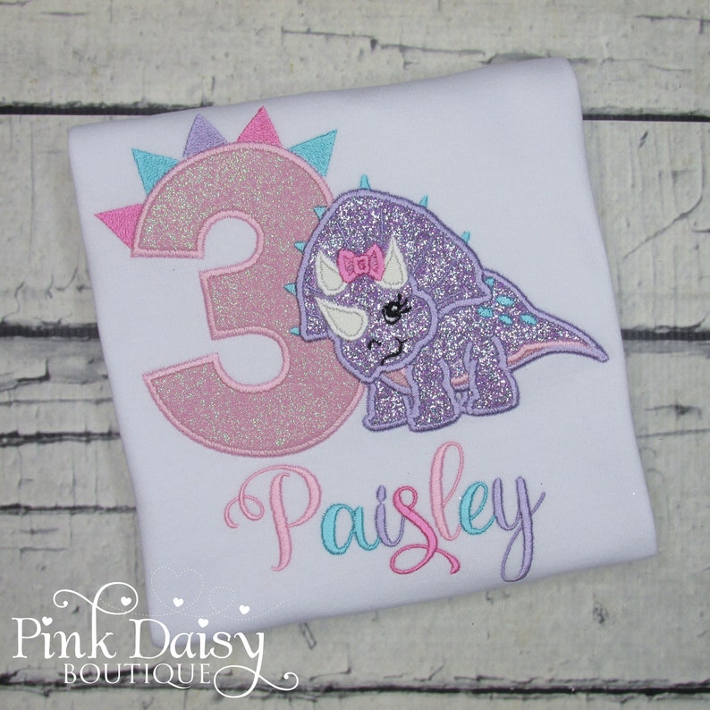 Puede incluir: Una camiseta blanca con un n&uacute;mero 3 brillante de color rosa y morado y un dinosaurio brillante de color morado con un lazo rosa. El nombre "Paisley" est&aacute; bordado en rosa, azul y morado.