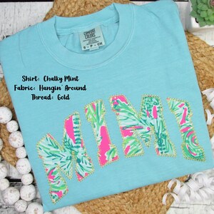 May include: A light blue t-shirt with the name "MIMI" appliqued in a tropical floral pattern with pink and green accents, outlined in gold thread. The shirt has a crew neck and a Comfort Colors tag.