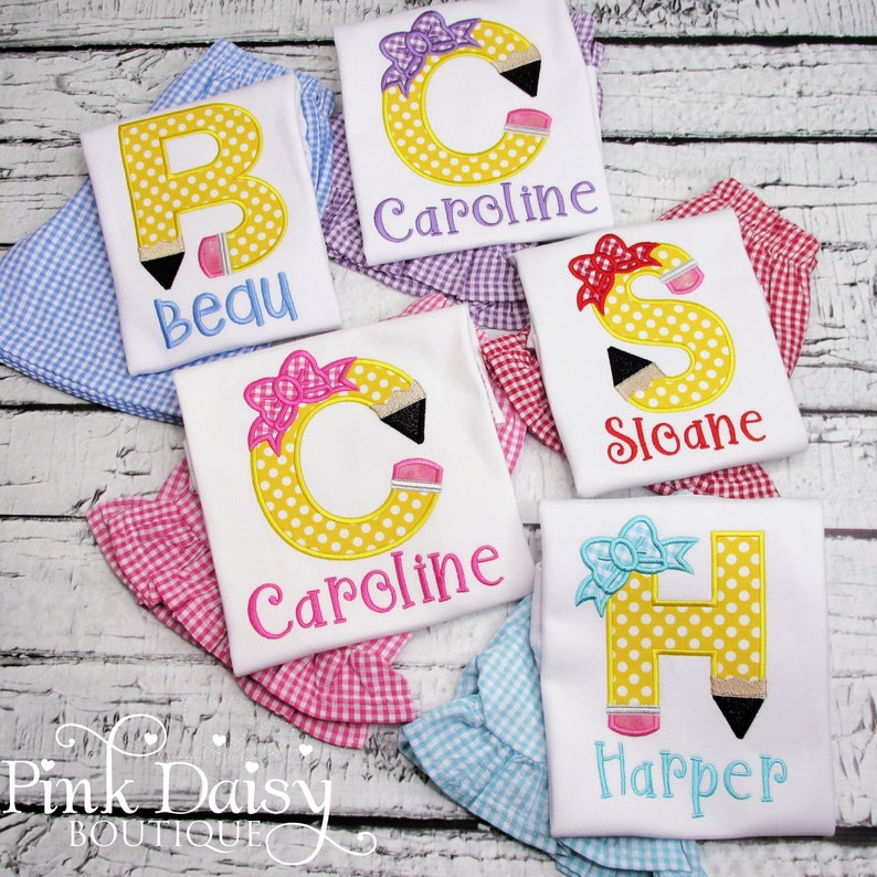 May include: Five white t-shirts with yellow polka dot letters that are shaped like pencils. Each shirt has a different letter and a name embroidered on the bottom. The names are Beau, Caroline, Sloane, and Harper.