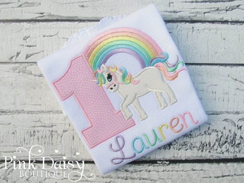 May include: White onesie with a pink number one with white polka dots, a rainbow, and a white unicorn with a rainbow mane and tail. The name "Lauren" is embroidered in rainbow colors below the unicorn.