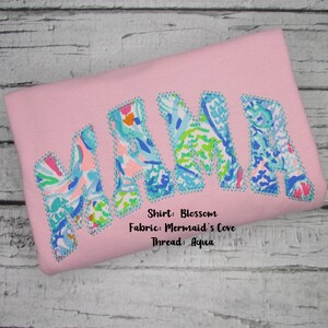 May include: Pink shirt with the word "MAMA" in large, colorful letters. The letters are filled with a floral pattern in shades of blue, green, and orange, outlined with aqua stitching. The shirt is made of Blossom fabric.