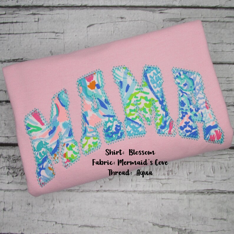 May include: Pink shirt with the word "MAMA" in large, colorful letters. The letters are filled with a floral pattern in shades of blue, green, and orange, outlined with aqua stitching. The shirt is made of Blossom fabric.