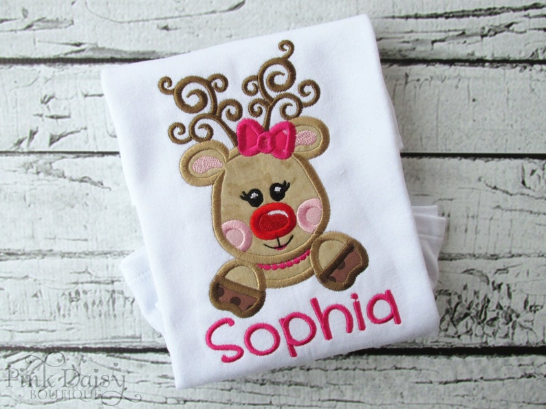 Puede incluir: Camiseta blanca con un reno marr&oacute;n bordado con mejillas rosadas, una nariz roja y un lazo rosa. El reno tiene cuernos marrones y est&aacute; sentado. El nombre "Sophia" est&aacute; bordado en rosa debajo del reno.