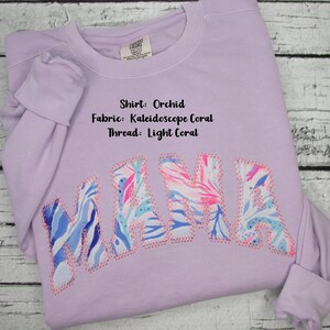 May include: Orchid-colored sweatshirt with the word "MAMA" appliqued across the front. The letters are made of Kaleidoscope Coral fabric and outlined with light coral thread. The shirt's details include the fabric and thread colors.