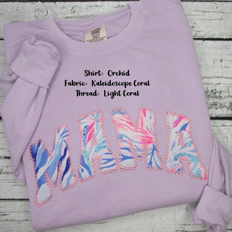 May include: Orchid-colored sweatshirt with the word "MAMA" appliqued across the front. The letters are made of Kaleidoscope Coral fabric and outlined with light coral thread. The shirt's details include the fabric and thread colors.