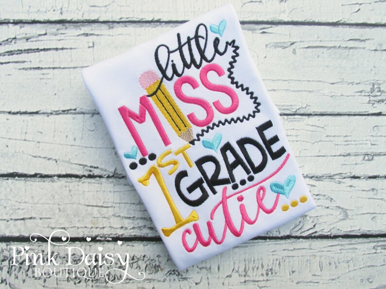 May include: White t-shirt with embroidered text that reads "little miss 1st grade cutie..." in pink, black, yellow, and blue. The text is surrounded by small hearts.