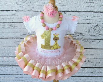 Puede incluir: Un tutú rosa y dorado con un body blanco que presenta un número uno de purpurina dorada y un gráfico de jirafa. El body está personalizado con el nombre "Gabriella".