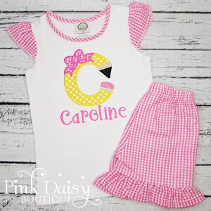May include: A white sleeveless shirt with a pink and white gingham trim and a yellow pencil-shaped letter C with pink polka dots and a pink bow. The shirt is personalized with the name "Caroline" in pink thread. The shirt is paired with pink and white gingham shorts.