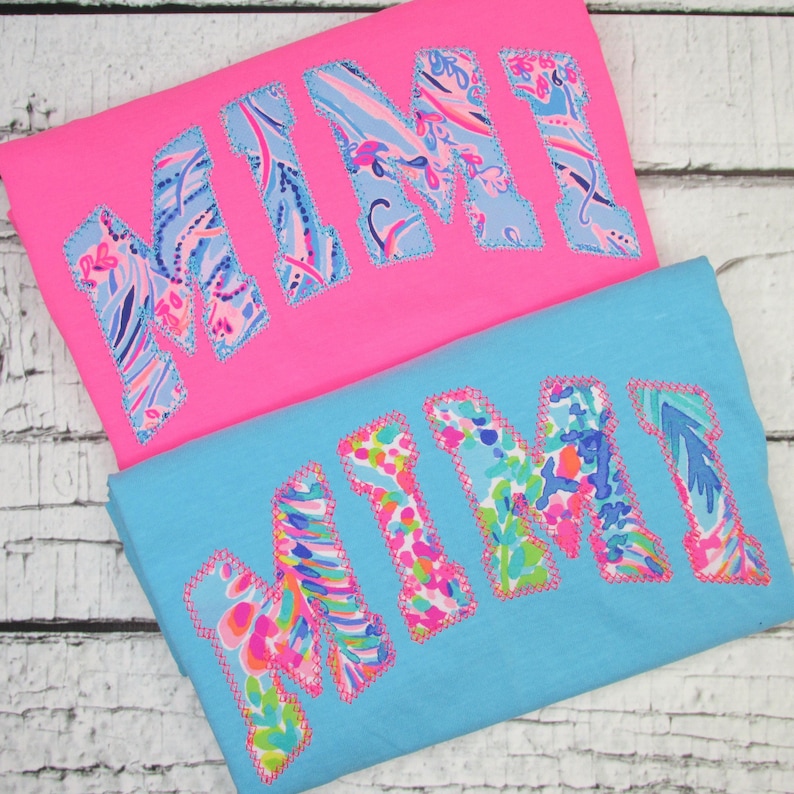 May include: Two t-shirts, one pink and one blue, each with the name "MIMI" appliqued in a colorful floral pattern. The letters are outlined with a contrasting stitch, creating a playful and eye-catching design. The shirts are folded and ready to wear.
