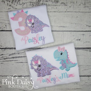 Puede incluir: Dos camisetas blancas con dinosaurios bordados y nombres. La camiseta de arriba tiene un n&uacute;mero 3 rosa y el nombre "Paisley". La camiseta de abajo tiene un dinosaurio morado y un dinosaurio azul con los nombres "Paisley's Mimi".