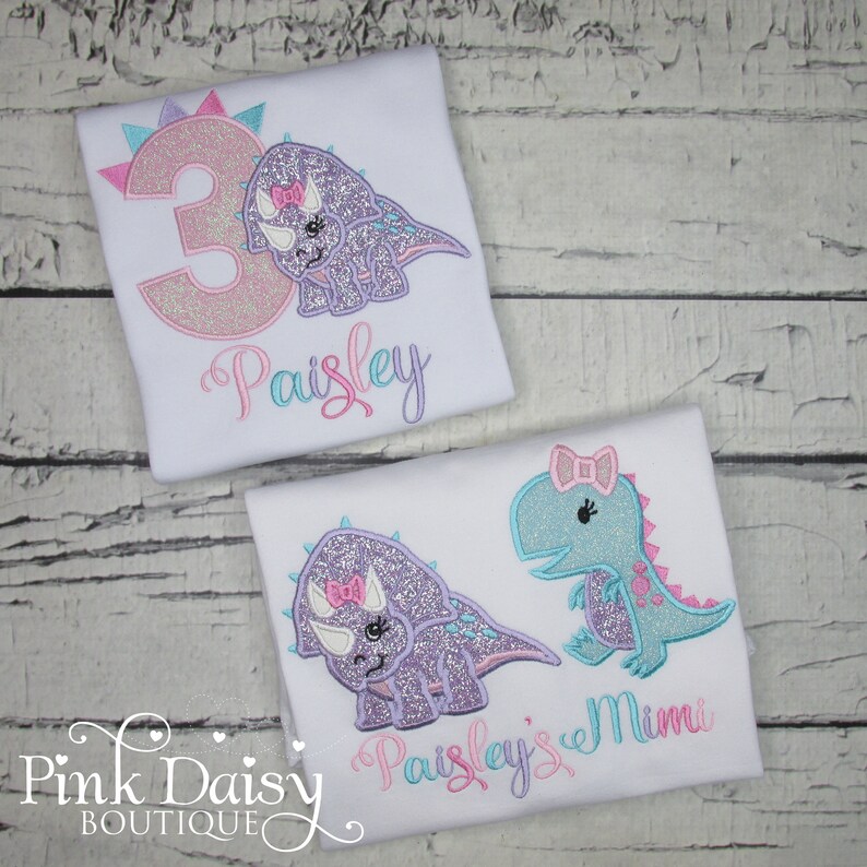 Puede incluir: Dos camisetas blancas con dinosaurios bordados y nombres. La camiseta de arriba tiene un n&uacute;mero 3 rosa y el nombre "Paisley". La camiseta de abajo tiene un dinosaurio morado y un dinosaurio azul con los nombres "Paisley's Mimi".