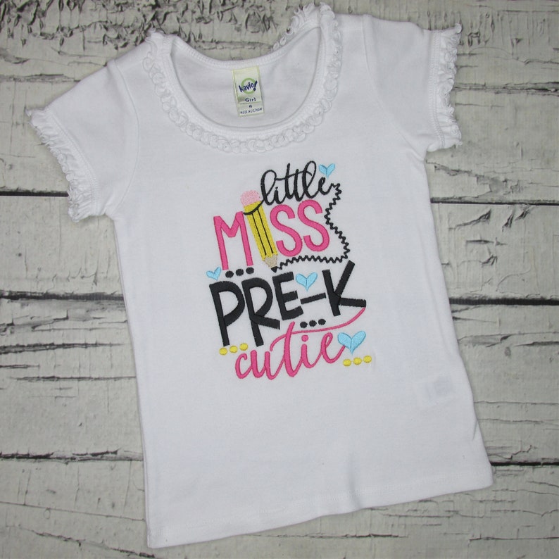 May include: White t-shirt with a pink, black, and yellow embroidered design that says "little miss pre-k cutie" with a pencil graphic.