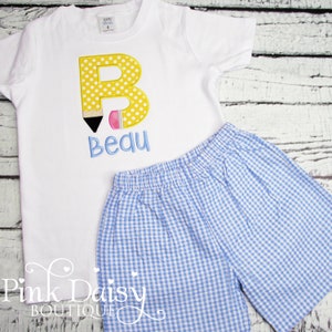 May include: A white short-sleeved shirt with a yellow letter B with polka dots, a black and pink pencil, and the name "Bequ" embroidered on it. The shirt is paired with blue and white gingham shorts.