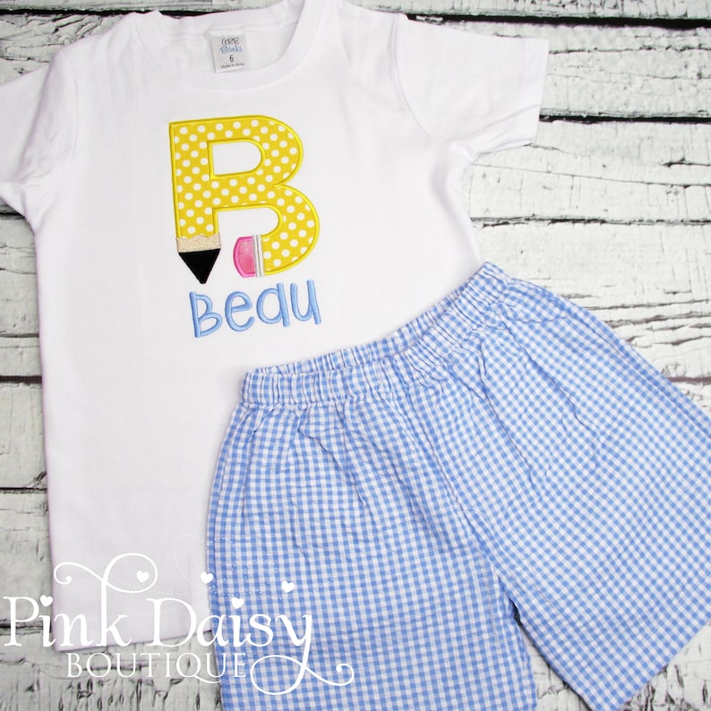 May include: A white short-sleeved shirt with a yellow letter B with polka dots, a black and pink pencil, and the name "Bequ" embroidered on it. The shirt is paired with blue and white gingham shorts.
