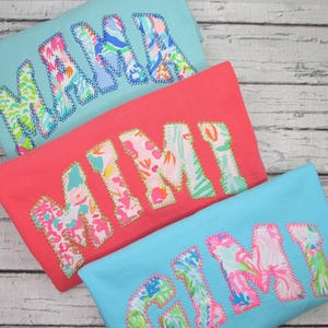 May include: Three colorful t-shirts with the words "MAMA" and "MIMI" appliqued on them. The letters are filled with a floral pattern and outlined with a contrasting stitch. The shirts are in shades of coral, light blue, and turquoise.