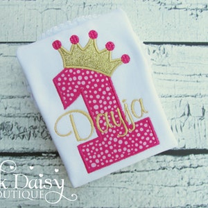 May include: White onesie with a pink and white polka dot number one and a gold glitter crown. The name "Daya" is embroidered in gold below the number one.