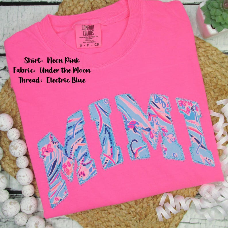 May include: Neon pink t-shirt with the name "MIMI" appliqued across the front in a blue and patterned fabric. The shirt has a crew neck and short sleeves. The shirt's fabric is "Under the Moon" and the thread is "Electric Blue".