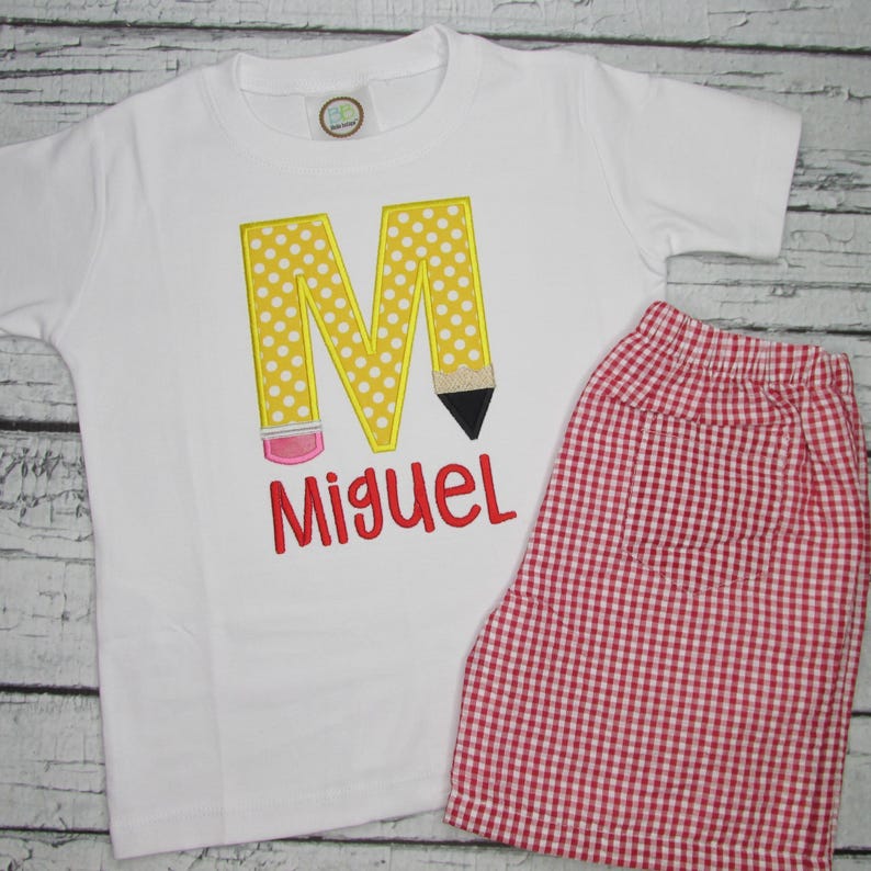 May include: A white t-shirt with a yellow and white polka dot letter M shaped like a pencil. The name Miguel is embroidered in red below the letter. The shirt is paired with red and white gingham shorts.