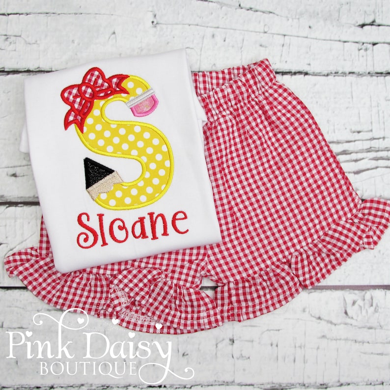 May include: A white t-shirt with a yellow letter S with polka dots and a red bow. The letter S has a black and white pencil behind it. The shirt says "Sloane" in red embroidery. The shirt is paired with red and white gingham shorts with a ruffle trim.
