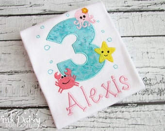 May include: White t-shirt with a turquoise number 3, decorated with an octopus, starfish, and crab. The name "Alexis" is embroidered in pink below the number. Bubbles are embroidered around the number.