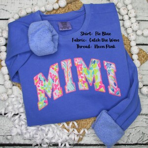 May include: Flo Blue sweatshirt with the name "MIMI" in a colorful floral pattern. The sweatshirt has a crew neck and long sleeves with cuffed ends. The text "Shirt: Flo Blue, Fabric: Catch the Wave, Thread: Neon Pink" is visible.