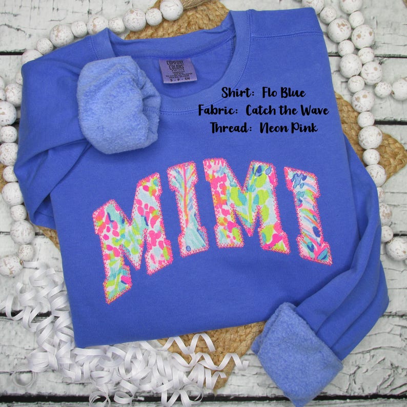 May include: Flo Blue sweatshirt with the name "MIMI" in a colorful floral pattern. The sweatshirt has a crew neck and long sleeves with cuffed ends. The text "Shirt: Flo Blue, Fabric: Catch the Wave, Thread: Neon Pink" is visible.