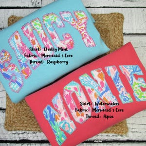May include: Two colorful shirts with embroidered names. One is light blue with the name "LUCY" in a floral pattern, and the other is red with the name "MONNIE". The shirts have text that reads "Shirt: Chalky Mint, Fabric: Mermaid's Cove, Thread: Raspberry" and "Shirt: Watermelon, Fabric: Mermaid's Cove, Thread: Aqua".