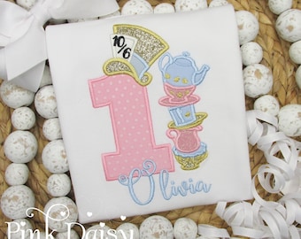 May include: A white t-shirt featuring an embroidered design of a large pink polka-dot number one, a gold glitter top hat with a 10/6 tag, and a stack of blue and pink teapots and cups. The name Olivia is embroidered in light blue script.