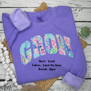 May include: A violet sweatshirt with the word "CROWN" appliqued across the front in a colorful floral pattern. The sweatshirt has a crew neck and long sleeves with a soft, fleece-lined interior. The text on the image says "Shirt: Violet, Fabric: Catch the Wave, Thread: Aqua."